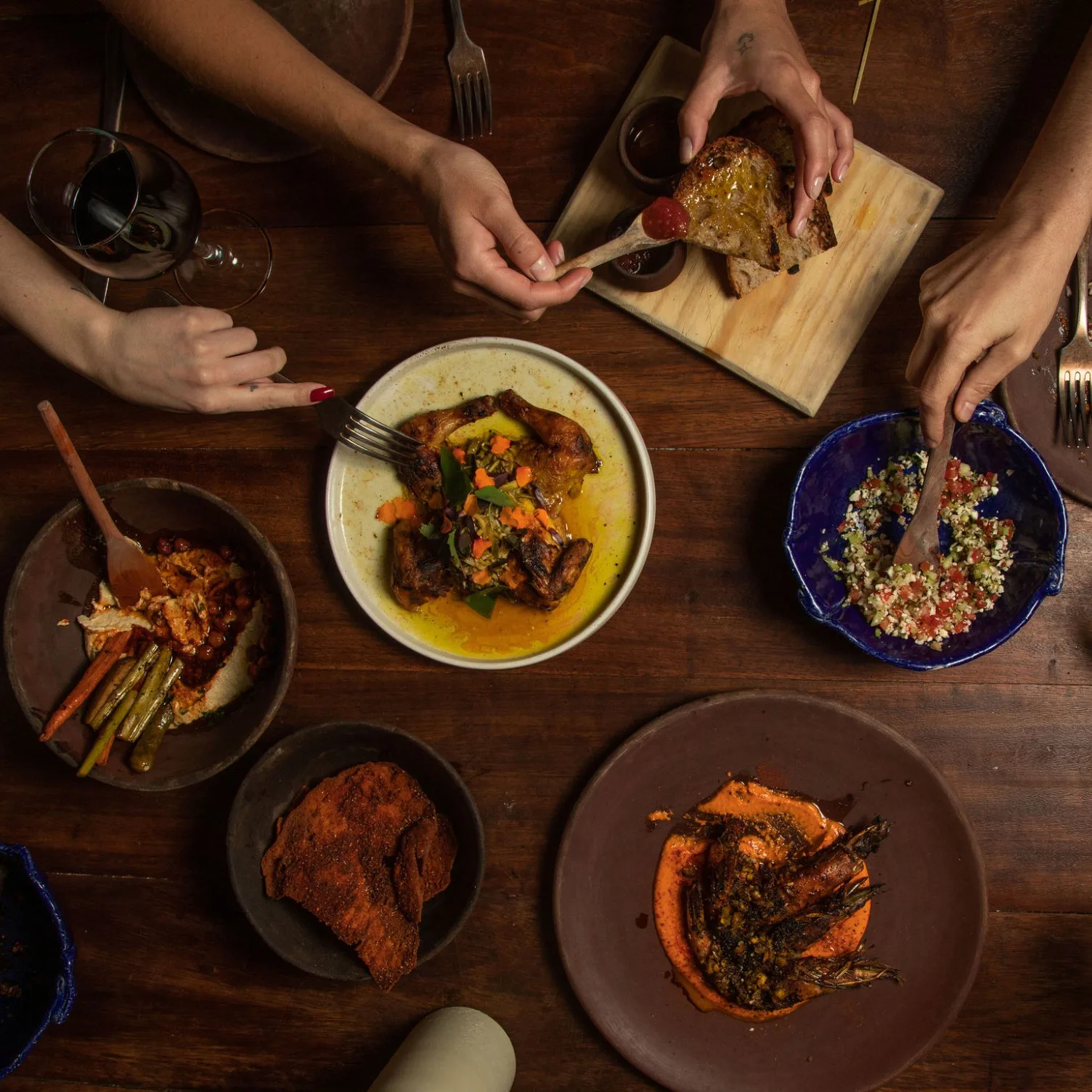 Mesa compartida con platos al centro, pan, vegetales, vino y manos sirviendo.