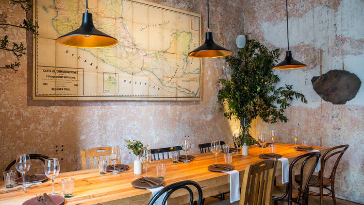 Mesa larga de madera junto a un muro con pátina y vegetación en el comedor.